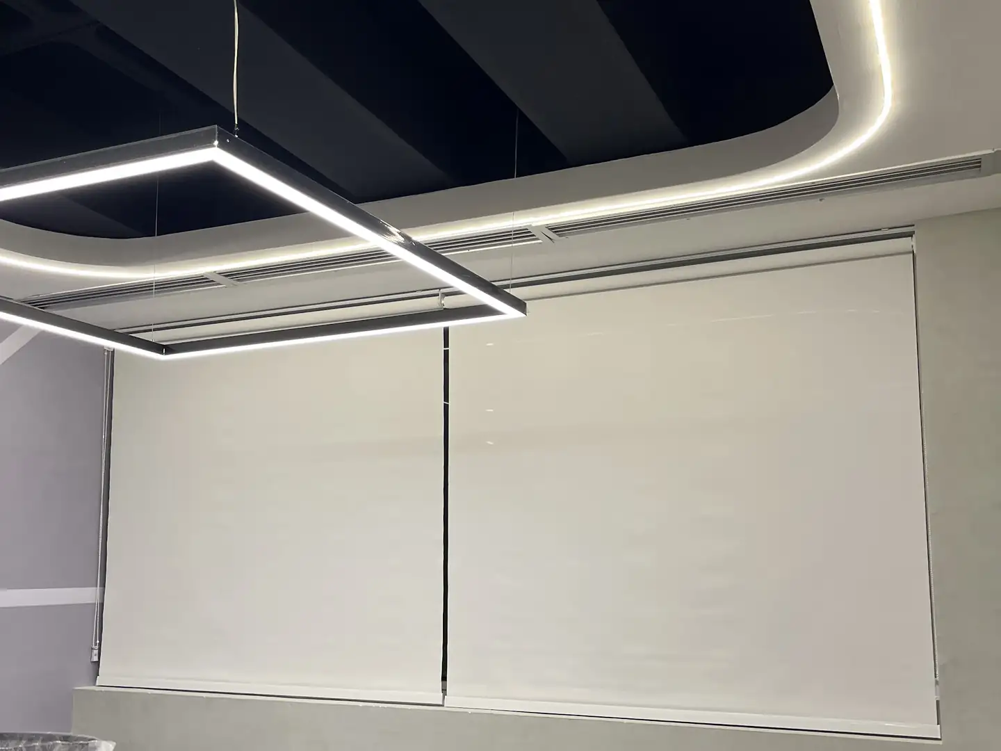 Duas telas solares em rolô em uma sala de reunião com luminária suspensa