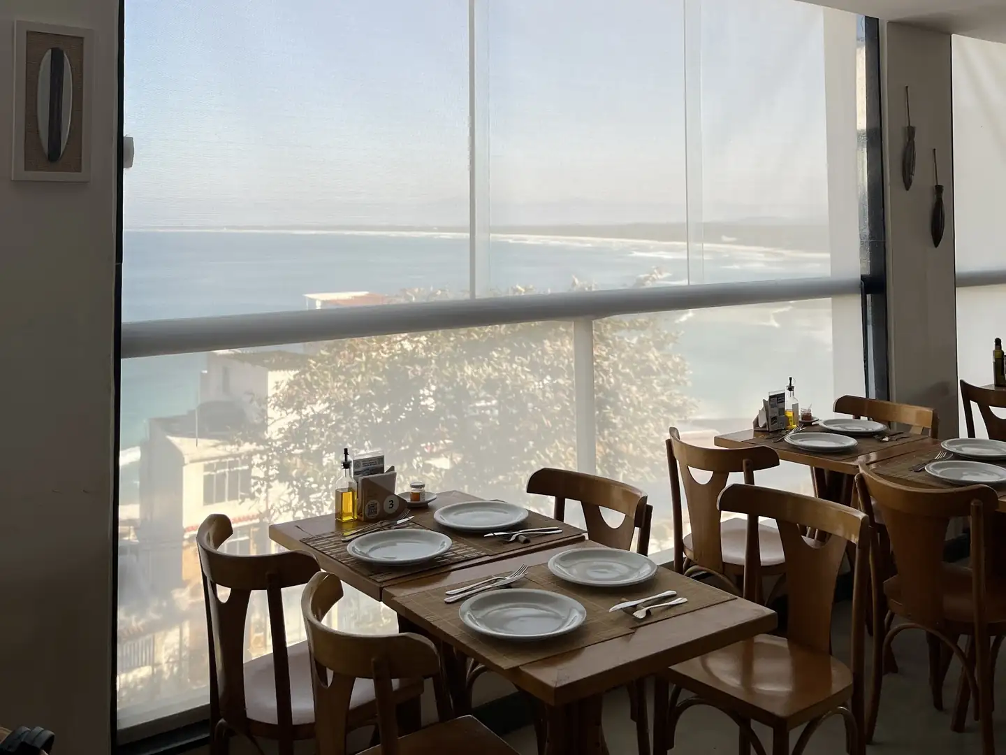 Mesas de um restaurante junto à parede de vidro com telas solares e o mar ao fundo